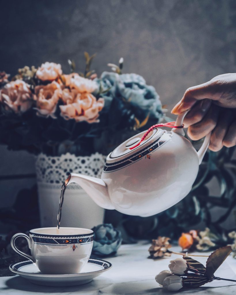 A hand holding a tilted teapot that is pouring English breakfast tea from Assam leaves into a tea cup resting on a saucer with a bouquet behind it.