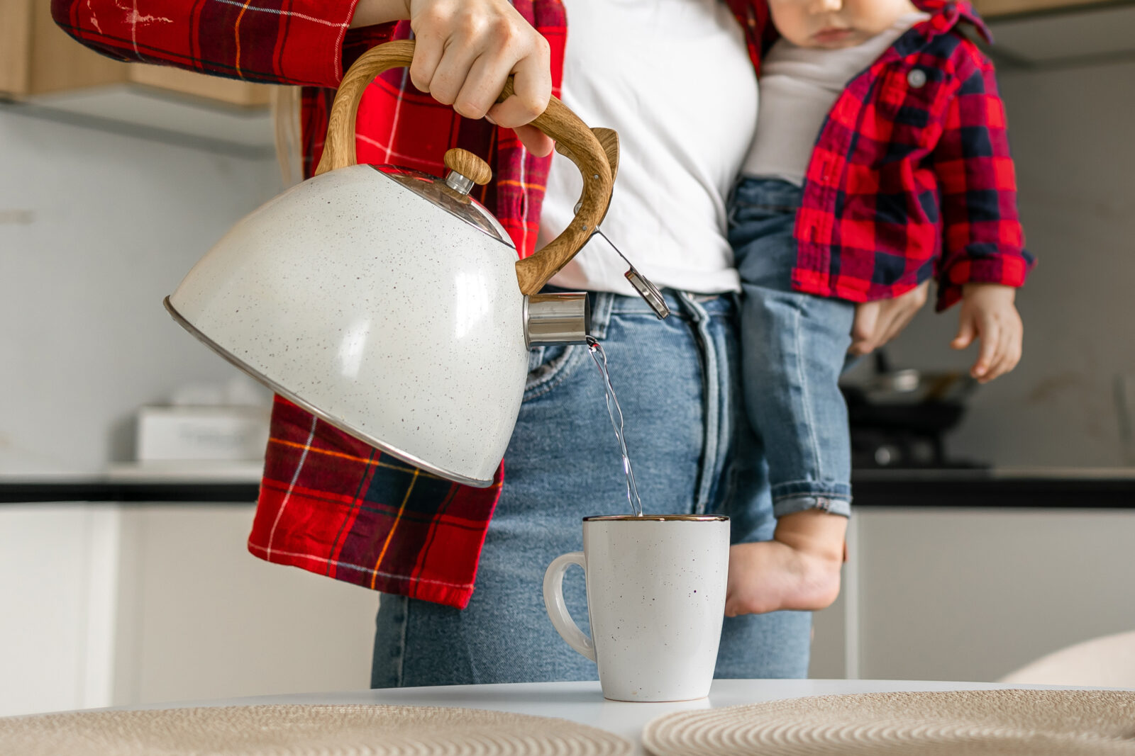 Smiling young woman pouring water from kettle into cup holding baby on hands